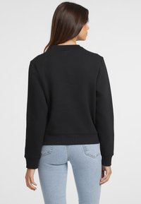 Felpa nera con colletto rotondo, polsini e orlo a costine, e una texture liscia. Abbinata a jeans blu chiaro.