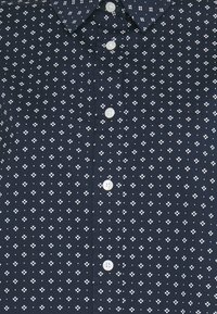 Chemise bleu marine boutonnée avec un motif floral blanc et à pois répétitifs, avec une poche poitrine côté droit.