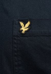 Κίτρινο κεντητό πουλί σε μπαλώμα στην επάνω γωνία μιας τσέπης από σκούρο ναυτικό ύφασμα, που δείχνει λεπτομερή ραφή και υφή υφάσματος.