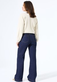 Cremefarbener, strukturierteter Pullover mit langen Ärmeln, kombiniert mit dunkelblauen Schlagjeans, die über Gesäßtaschen und kontrastierende Nähte verfügen.