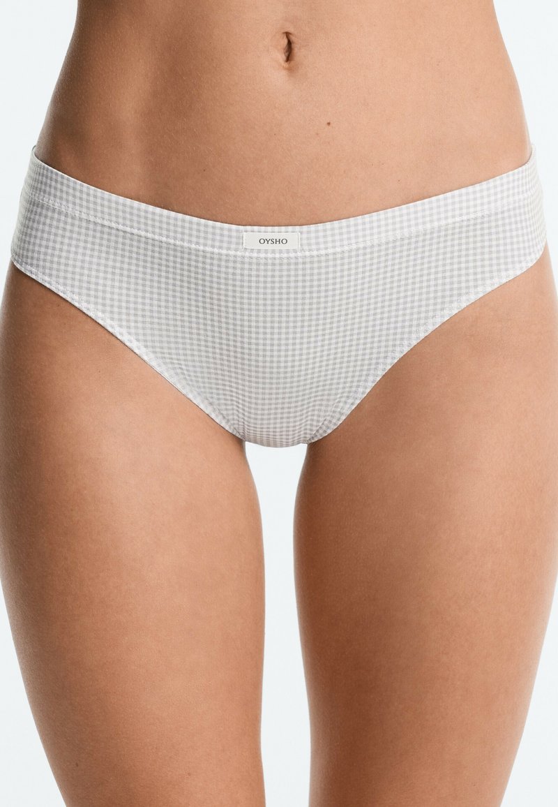 Gros plan sur la partie basse du torse d’une femme portant un sous-vêtement style bikini à motif vichy gris clair et blanc avec une petite étiquette "OYSHO" sur la ceinture.