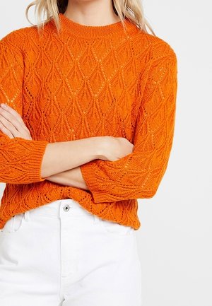 Femme les bras croisés, portant un pull en maille texturée orange vif et un pantalon blanc taille haute sur un fond uni.