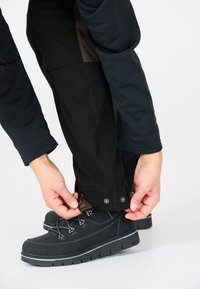 Zwarte outdoorbroeken met verstelbare boorden, die worden aangetrokken. Een langemouwen donker shirt en stevige zwarte wandelschoenen zijn ook zichtbaar.