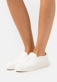 Des baskets en toile blanches présentent un design noué à l'avant, une semelle en caoutchouc et une texture lisse. La tige a une forme basse et est facile à enfiler.