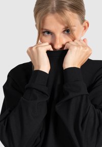 Zwarte longsleeve shirt met hoge kraag, licht oversized pasvorm en zachte textuur, omhoog gehouden tegen het gezicht. Eenvoudig ontwerp zonder zichtbare patronen.