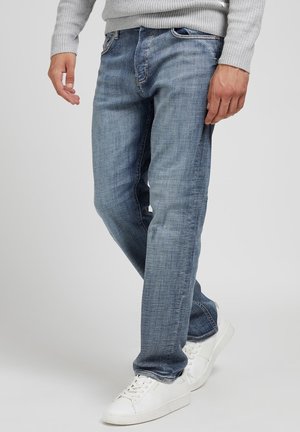 Mężczyzna ubrany w jasnoniebieskie dżinsy, białe trampki i jasnoszary, prążkowany sweter, stojący z jedną lekko ugiętą nogą.