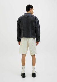 Veste en denim noir, coupe oversize, coutures visibles, manches courtes. Shorts blancs avec poches cargo, montrés de dos. Chaussures et chaussettes noires.