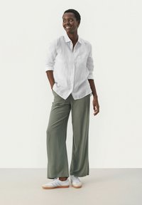 Chemise blanche en lin à boutons, coupe décontractée, associée à un pantalon large vert olive et des baskets blanches à semelles en gomme, sur fond neutre.
