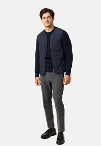 Rakenteellinen tummansininen liivi, jossa on tikkikuvio, sivutaskut, käytetty tumman neuleen ja harmaiden housujen päällä. Mustat kengät täydentävät asua.