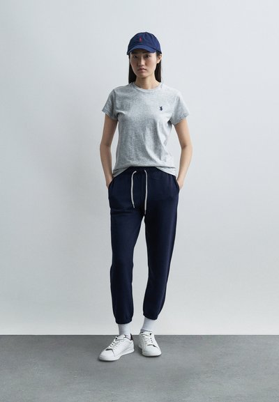 Γυναίκα που στέκεται με τα χέρια στις τσέπες φορώντας γκρι μπλουζάκι, σκούρο μπλε φόρμα, λευκά αθλητικά παπούτσια, λευκές κάλτσες και σκούφο μπέιζμπολ σε σκούρο μπλε χρώμα.