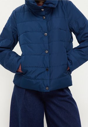 Veste matelassée bleue avec boutons-pression portée sur un pantalon bleu foncé, mains dans les poches latérales, debout devant un fond uni.