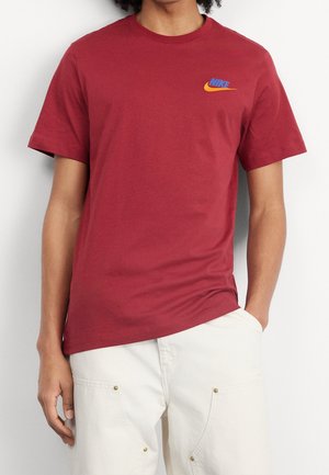Persona que lleva una camiseta roja de Nike con un pequeño logo azul y naranja en el pecho y pantalones claros con bolsillos.