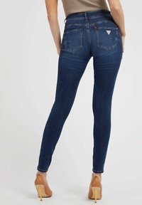 Kvinna i mörkblå skinny jeans med bakfickor och tanfärgade högklackade skor, stående mot en enfärgad ljus bakgrund.