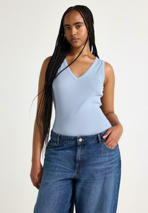 Ung kvinde med langt flettet hår iført en lyseblå ærmeløs V-hals top og løse blå denimjeans med den ene hånd i lommen.
