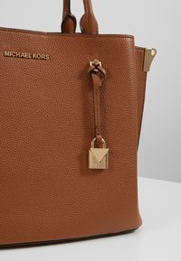 Brązowa skórzana torebka o teksturowanym wykończeniu, z złotymi akcentami, z zamkiem w kształcie kłódki i wytłoczonym logo marki z przodu.