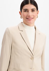 Blazer beige en tissu texturé, fermeture à un bouton, et revers crantés. Porté par-dessus un col roulé blanc à côtes. Boucles d'oreilles en or en forme de créole.