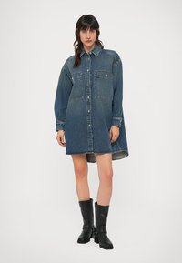 Φόρεμα πουκάμισο από τζιν με κουμπιά στο εμπρός μέρος, δύο τσέπες στο στήθος και μακριά μανίκια. Διαθέτει φαρδιά γραμμή και κυρτό τελείωμα. Σκούρο μπλε χρώμα.