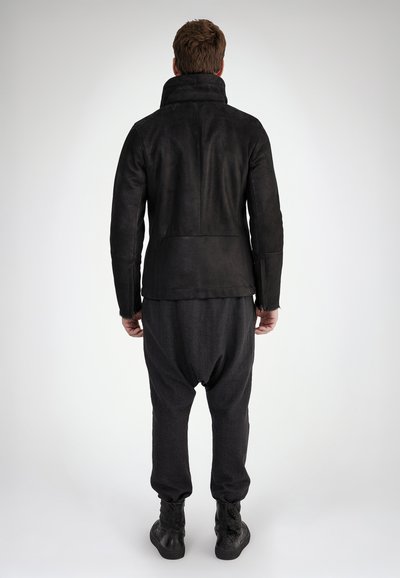 Schwarze Wildlederjacke mit hohem Kragen, kombiniert mit dunklen, eng zulaufenden Hosen im Drop-Crotch-Design und schwarzen Schnürstiefeln.