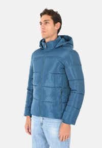 Giacca imbottita con cappuccio di un profondo blu. Presenta una finitura lucida, cuciture verticali e tasche laterali con zip. Design leggero.