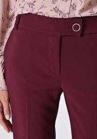Bordeauxkleurige broek met een gladde textuur, voorzien van een platte voorkant, zijzakken en een decoratieve accent van zilverkleurig beslag bij de tailleband.