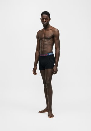 Mustan meessoost modell seisab paljajalu, kandmas musti bokserid, mille vöökohal on sinine ja punane Björn Borgi riba, lihtsa valge tausta ees.