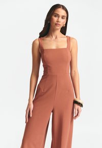 Rostfärgad jumpsuit med fyrkantig halsringning, breda axelremmar och avslappnad passform. Slät textur, med sidofickor och skräddarsydda sömdetaljer.