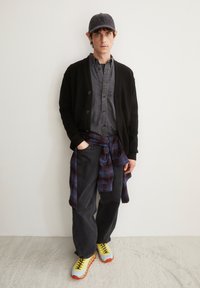 Schwarze Strickjacke, dunkles Hemd, navy Karoshirt um die Taille gebunden, dunkle Hose und leuchtend gelbe Sneaker mit orangefarbenen Akzenten.