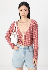 Top court à carreaux rouges et blancs avec des manches longues, un décolleté en V profond, une fermeture à nouer à l'avant et une garniture décorative. Associé à un short en denim bleu clair.