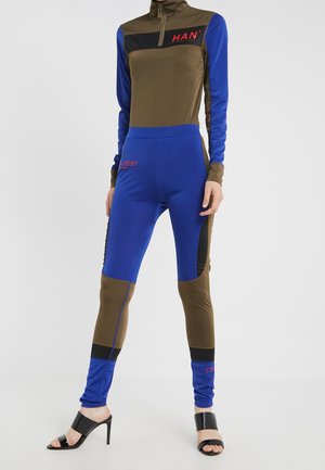 Femme portant un haut zippé à blocs de couleurs bleu et olive et un legging assorti, avec des sandales à talons hauts noires, debout devant un fond blanc.