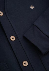 Granatowy sweter zapinany na guziki o gładkiej fakturze, z drewnianymi guzikami, haftowanym emblematem i dekoltem w kształcie litery V.