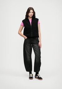 Svart fleecevest med glidelås foran, rosa kortermet skjorte under, mørke baggy jeans og svarte joggesko med gummisåler.