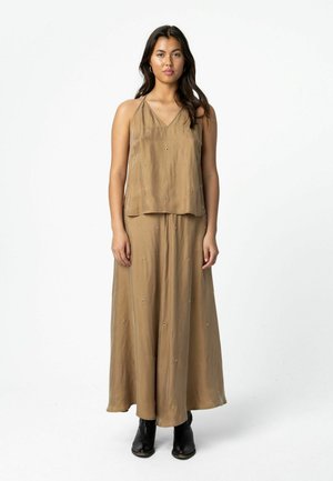 Femme portant un haut marron clair sans manches à col en V et un pantalon large assorti, avec des bottines noires, debout devant un fond blanc uni.