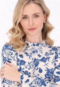 Blusa floreale a maniche lunghe con motivi di rose blu e farfalle su uno sfondo crema. Presenta un collo arricciato e maniche a sbuffo.