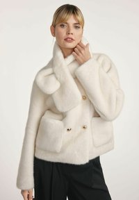 Kobieta z blond włosami ubrana w białą, puszystą kurtkę dwurzędową z dużymi kieszeniami i szerokim kołnierzem, w parze z czarnymi spodniami.