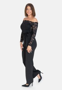 Top in pizzo nero a spalle scoperte con maniche lunghe, abbinato a pantaloni neri a vita alta e tacchi a punta neri. Tessuto morbido e dettaglio a fiocco.