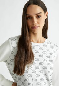 Pullover bianco a maglia con motivo circolare grigio e dettagli con logo. Presenta maniche lunghe in tessuto bianco uniforme. Texture liscia, design aderente.