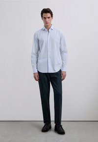 Camisa azul celeste de botones con mangas largas, combinada con pantalones verde oscuro. La camisa presenta un cuello clásico y un dobladillo recto.