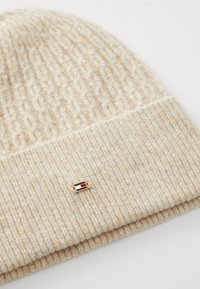 Strikket beige lue med et teksturert mønster, med en liten gullhardwaredetalj og et rødt, hvitt og blått logo på forsiden.