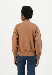 Sudadera marrón con escote redondeado, dobladillo acanalado y corte holgado. Combinada con vaqueros de mezclilla azul, mostrando un estilo relajado.