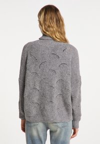 Grijze gebreide cardigan met een textuurpatroon in golven, geribbelde manchetten en een sjawl-kraag. Draagt over lichtblauwe denim jeans.