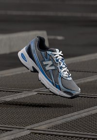 Bėgimo batelis su juodu tinklelio viršumi, pilkais ir mėlynais akcentais, baltais raišteliais ir storu baltu padu su švelniu išlinkimu.