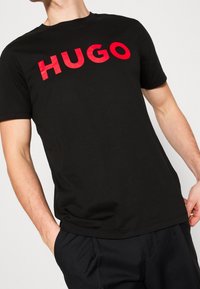 Schwarzes T-Shirt aus Baumwolle mit einem großen roten "HUGO"-Logo auf der Vorderseite. Kurze Ärmel und ein klassisches Rundhalsausschnitt-Design.