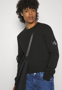 Jauns vīrietis ar cirtainiem matiem, uzvilcis melnu garām piedurknēm džemperi ar CK logotipu uz piedurknes, zilu džinsus un melnu plecu somu.