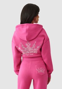 Różowa bluza z kapturem z designem korony i dżetami na plecach. Połączona z dopasowanymi spodniami, które mają mniejszy detal korony na kieszeni.