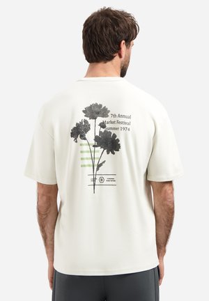 Man wearing a white T-shirt with black floral print and text "7th Annual Market Festival Summer 1974" on the back, standing facing away.