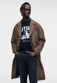 Καφέ παλτό από μίγμα μαλλιού, σχεδιασμένο με γραμμή και γιακάδες με σχισμή. Φορεμένο πάνω από σκούρο μπλε πουλόβερ με γραφικό βουνό και σκούρο τζιν. Ορατή μαύρη ζώνη.