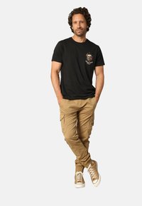 Zwarte graphic t-shirt met een bloemskullontwerp, gecombineerd met khaki cargobroeken en tan sneakers met witte zolen.