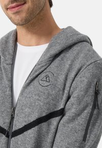 Grå zip-up hoodie med mjuk textur, svart randmönster och en broderad logotyp på bröstet. Har zip-fickor på sidorna.