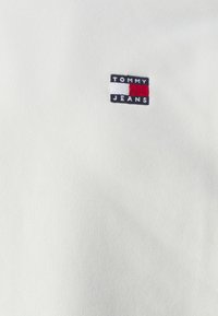 Bílá bavlněná látka se malým obdélníkovým logem s nápisem "TOMMY JEANS" v černé barvě, lemovaným červenými a bílými pruhy.