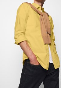 Man som bär en gul skjorta med knappar och uppkavlade ärmar, en beige tröja draperad över axlarna, samt mörka jeans med ena handen i fickan.